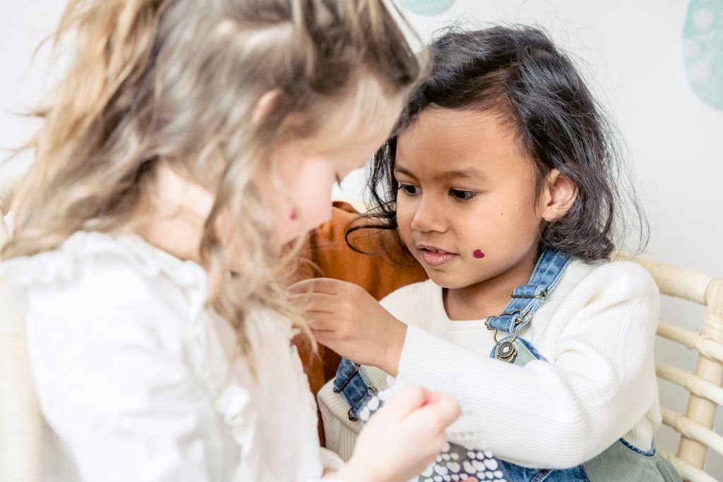 Concentrated multiracial preschool girls sticking colorful heart shaped stickers on each other while sitting near wall with egg decorations during Easter holiday